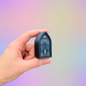 May include: A small, dark blue, arched window-shaped object with a stained-glass design. The window features a light blue background with yellow and gold accents. The object is held in a hand against a soft, colorful background.