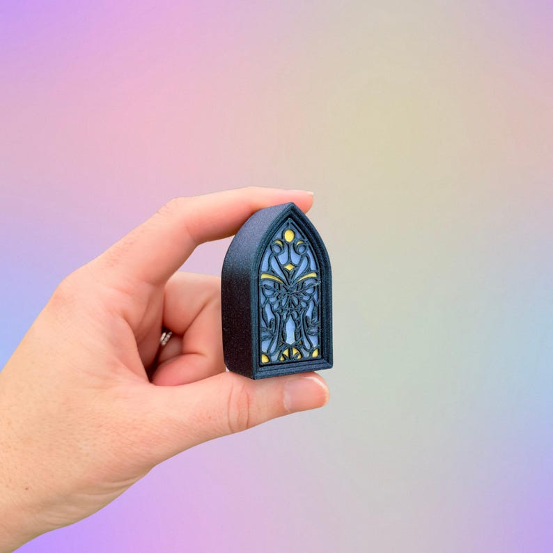 May include: A small, dark blue, arched window-shaped object with a stained-glass design. The window features a light blue background with yellow and gold accents. The object is held in a hand against a soft, colorful background.