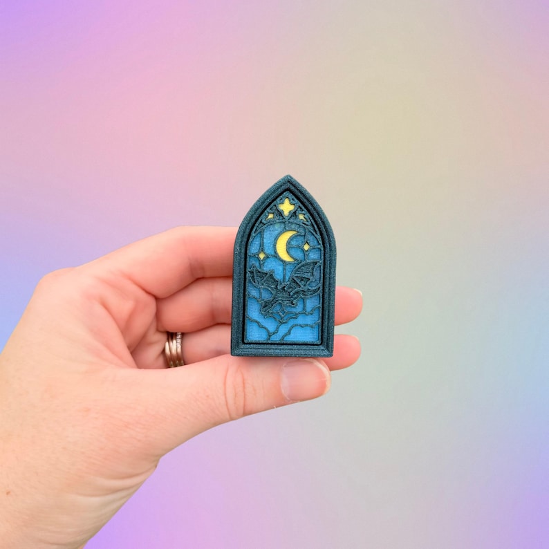 May include: A dark blue, arched-shaped pin featuring a night sky design. The pin has a yellow crescent moon, stars, and a bat silhouette. The background is a gradient of blue and yellow.