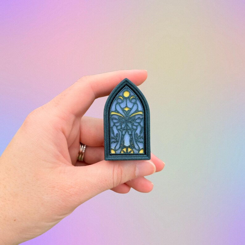 May include: A small, dark teal-colored, arched window-shaped object with a stained-glass design. The design features blue, yellow, and white elements. The object is held in a hand against a soft, pastel-colored background.