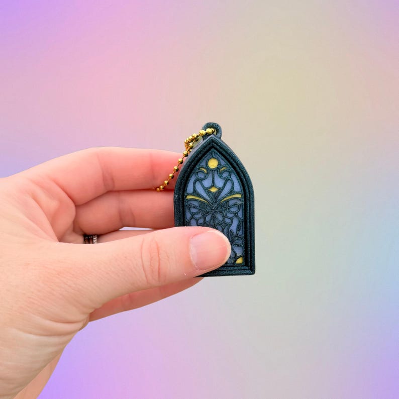May include: A small, arched keychain featuring a stained-glass window design. The window has a blue background with yellow and dark blue accents. A gold-colored chain is attached to the top. The keychain is held in a hand against a soft, colorful background.
