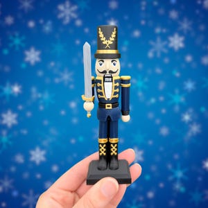 May include: A nutcracker figurine wearing a dark blue uniform with gold trim, a black hat with a gold laurel emblem, and black boots. The figure holds a silver sword in its right hand and stands on a square black base against a blue background.