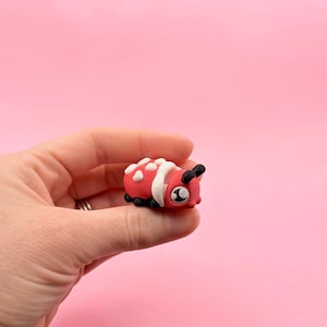 May include: A small, red and white ladybug figurine with black accents. The ladybug has white spots on its back and black antennae. The figurine is held in a hand against a pink background.