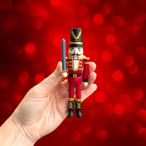 May include: A miniature wooden nutcracker soldier figurine, featuring a red uniform with gold accents, a black hat with a gold band and a sword. The toy soldier has a white mustache and is holding a gray sword. The background is a blurred red.