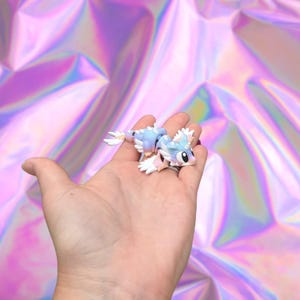 May include: A small, light blue and white fantasy creature figurine with a whimsical design. The figurine has large eyes and is held in a hand against a holographic background. The creature has a light blue body, white accents, and pink details.