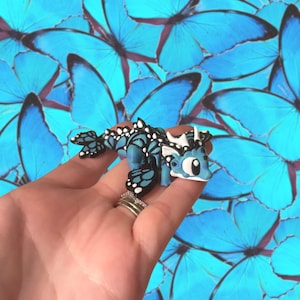 May include: A small, blue and black dragon figurine with white accents. The dragon has butterfly wing-like patterns and is held in a hand. The background is a pattern of blue butterfly wings.