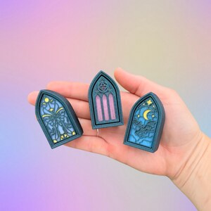 May include: Three teal-colored, arched window-shaped decorative pieces. Each piece features a different design: a butterfly, vertical stripes, and a moon with bats. The pieces are held in a hand against a pastel background.
