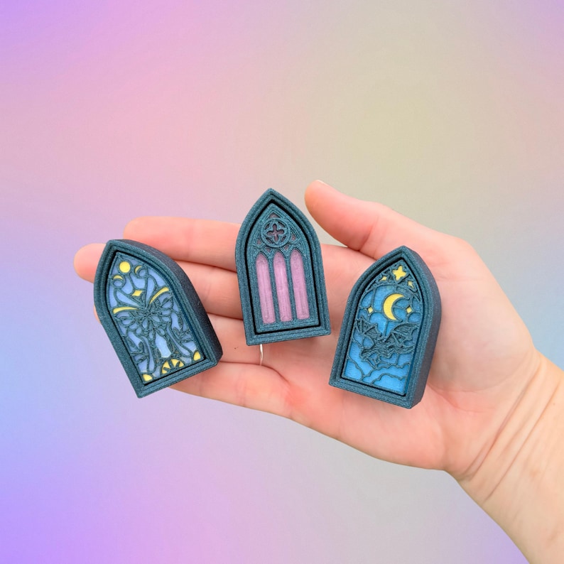 May include: Three teal-colored, arched window-shaped decorative pieces. Each piece features a different design: a butterfly, vertical stripes, and a moon with bats. The pieces are held in a hand against a pastel background.