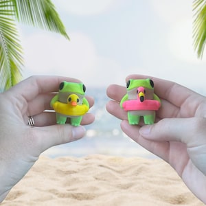 May include: Two green frog figurines, each with black eyes, are held up against a beach backdrop. One frog wears a yellow duck floatie, the other a pink flamingo floatie. The frogs are a fun summer theme.