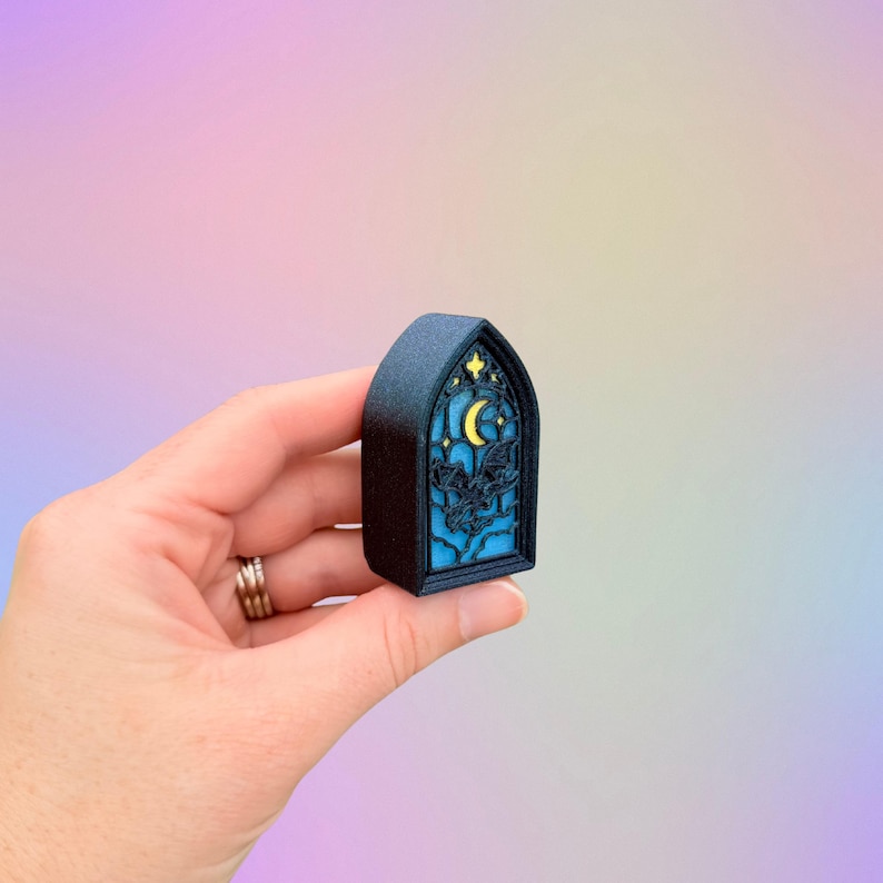 May include: A small, dark blue, arched decorative piece resembling a stained glass window. The design features a yellow crescent moon, stars, and silhouettes against a blue background. The piece is held in a hand.