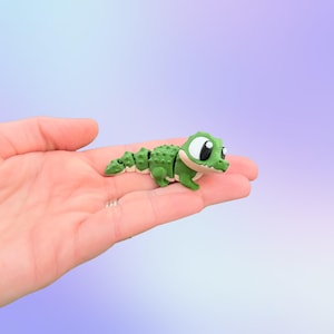 May include: A small, green alligator figurine with large, cartoonish eyes and a segmented body. The alligator is resting on a person's palm, showcasing its size and detail. The figurine has a textured back and a white underbelly.