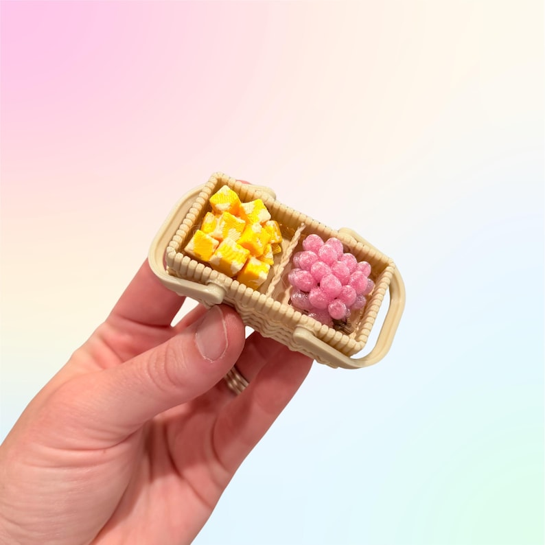 Puede incluir: Una cesta de picnic beige en miniatura con dos compartimentos. Un lado contiene alimentos c&uacute;bicos amarillos y blancos, el otro tiene un grupo de esferas rosas. La cesta tiene un asa y se sostiene en una mano.