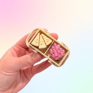 Puede incluir: Una cesta de picnic en miniatura con tres cu&ntilde;as de queso y un racimo de uvas rosas. La cesta es beige con un borde de cuentas y compartimentos divididos. Los alimentos son detallados.