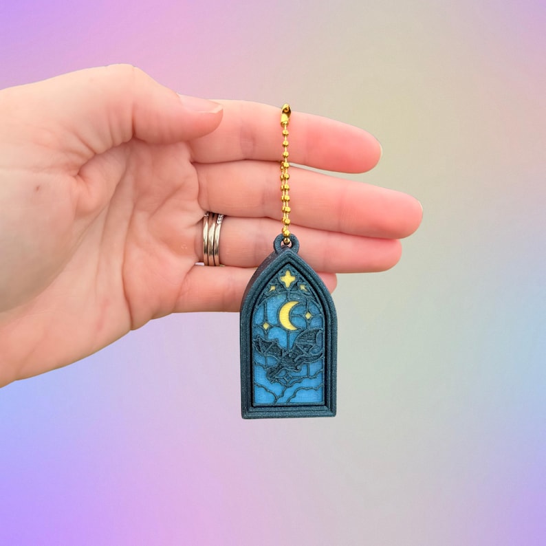 May include: A dark blue, arched pendant with a gold chain, featuring a night sky design. The pendant showcases a crescent moon, stars, and a dragon silhouette against a blue background. The pendant is held by a hand.