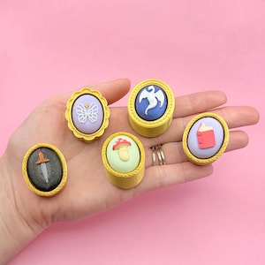 May include: A hand displays six colorful, round decorative objects. Each object has a gold-colored border and features a different design: a butterfly, a dragon, a mushroom, a book, and a sword. The objects are set against a pink background.