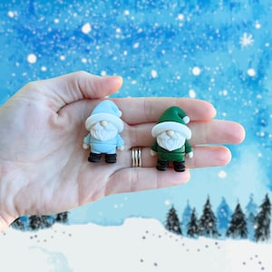 May include: Two miniature gnome figurines, one light blue and one green, each with a white beard and hat. The gnomes are held in a hand against a winter-themed backdrop with a snowy landscape and falling snowflakes.