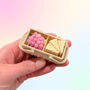 Puede incluir: Una peque&ntilde;a cesta de picnic con un racimo de uvas rosas y tres cu&ntilde;as de queso. La cesta es beige con un dise&ntilde;o tejido y un asa. Los alimentos son detallados y realistas, perfectos para casas de mu&ntilde;ecas o exhibiciones en miniatura.