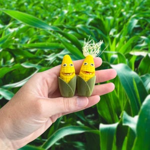May include: Two smiling corn-shaped figurines with yellow kernels and green husks. One has a small tuft of white hair. The background is a field of green corn stalks. The figurines are held in a hand.