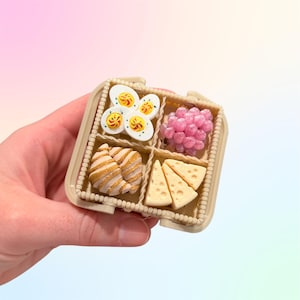 Puede incluir: Una peque&ntilde;a caja de comida con cuatro compartimentos, que contiene r&eacute;plicas en miniatura de huevos en rodajas, uvas rosas, cruasanes y cu&ntilde;as de queso. La caja es beige y se sostiene en una mano sobre un fondo suave y colorido.