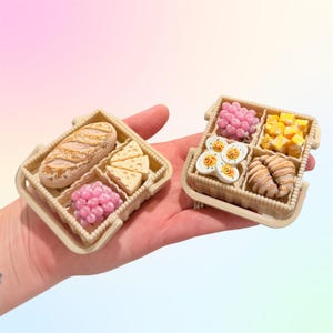 Puede incluir: Dos cestas de picnic en miniatura, cada una con una selecci&oacute;n de alimentos. Una cesta tiene un pan, cu&ntilde;as de queso y bayas rosas. La otra contiene bayas rosas, cubos amarillos, huevos en rodajas y cruasanes. Las cestas son beige.