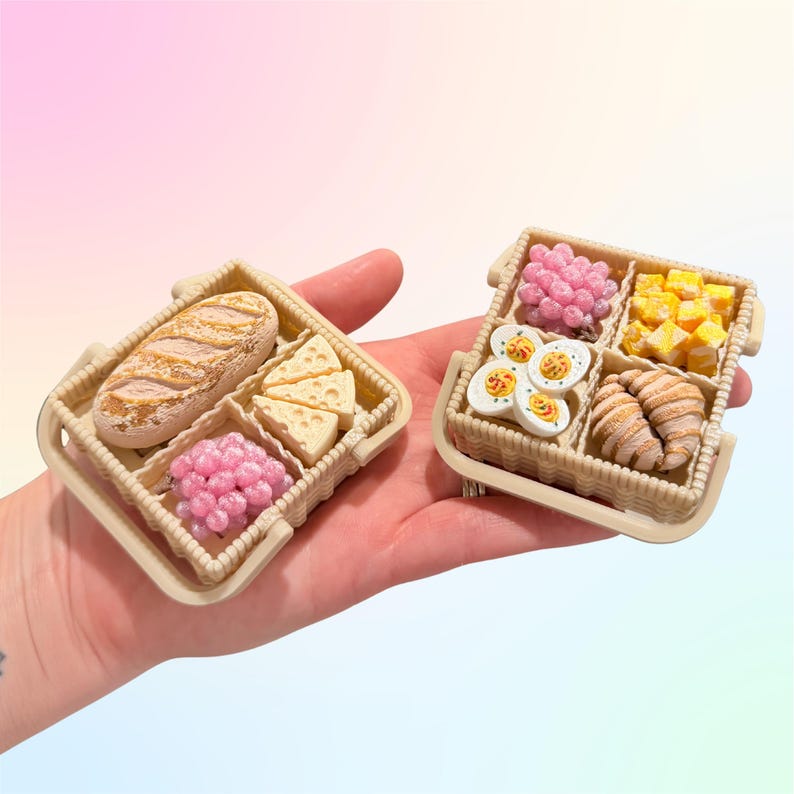 Puede incluir: Dos cestas de picnic en miniatura, cada una con una selecci&oacute;n de alimentos. Una cesta tiene un pan, cu&ntilde;as de queso y bayas rosas. La otra contiene bayas rosas, cubos amarillos, huevos en rodajas y cruasanes. Las cestas son beige.