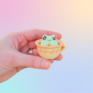May include: A small, light green frog figurine with large dark eyes and a smiling mouth, sitting inside a miniature cream-colored teacup. The teacup features a delicate, embossed orange leaf pattern around its exterior and a small handle.