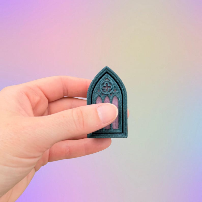 May include: A teal-colored, arched window miniature with a detailed cross design at the top. The window's interior is a soft pink, and the frame has a textured appearance. The miniature is held against a pastel gradient background.