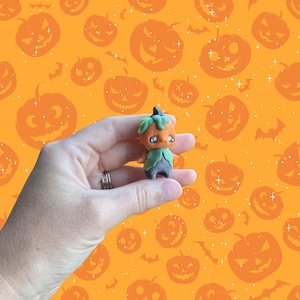 May include: A small, orange pumpkin-shaped figurine with a green leafy collar and a dark blue stem. The figurine has large, expressive eyes and is held against a background of orange pumpkins and bats.