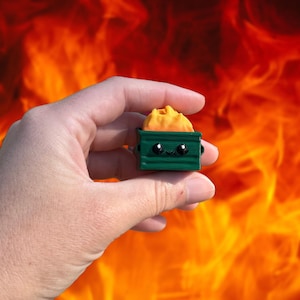 May include: A small, green dumpster figurine with a smiling face and a pile of yellow material inside. The figurine is held in a hand against a fiery orange and red background.