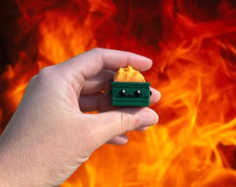 May include: A small, green dumpster figurine with a smiling face and a pile of yellow material inside. The figurine is held in a hand against a fiery orange and red background.