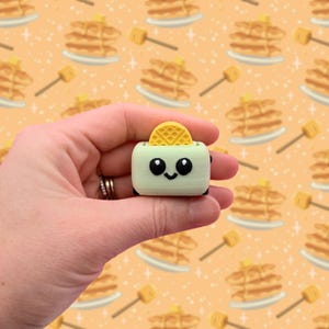 May include: A light green, cartoon-style toaster with a yellow waffle popping out. The toaster has a smiling face with large black eyes. The background is a repeating pattern of pancakes and honey dippers.