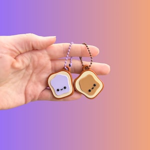 May include: Two toast-shaped keychains with smiling faces. One features a light purple spread with a purple ball chain, and the other features a light brown spread with a brown ball chain. Both have tan crusts and dark brown outlines.
