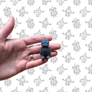 May include: A small, gray robot figurine with blue eyes. The robot has a rounded head, a rectangular body, and short legs. The background features a repeating pattern of gray robot illustrations on a white surface.