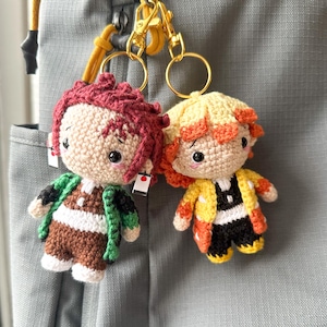 May include: Two crocheted anime character keychains. One has burgundy hair and a green and brown outfit. The other has yellow hair and a yellow, orange, and black outfit. Both have gold key rings and are attached to a gray backpack.