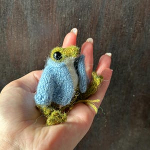 May include: A miniature, handmade frog ornament. The frog is predominantly green and white, wearing a light blue sweater. It has large black eyes and is crafted from yarn. This is a whimsical, handcrafted item.