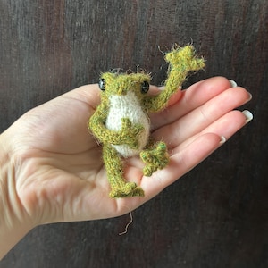 May include: A handmade, knitted frog figurine. The frog is primarily green with a white belly. It has black eyes and is posed with one arm raised. The frog is approximately 3 inches tall.