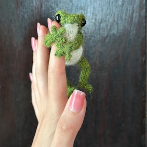 May include: A handmade, green and white knitted frog figurine with black eyes. The frog is perched on a finger, showcasing its detailed craftsmanship. The frog's body is a mix of green and white yarn, creating a textured appearance.