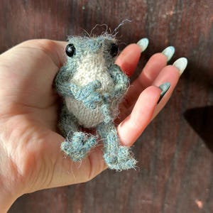 May include: A small, handmade frog figurine. The frog is crafted from blue and white yarn, with black bead eyes. The frog is posed with its legs extended, giving it a lifelike appearance. The frog is a whimsical, handcrafted item.