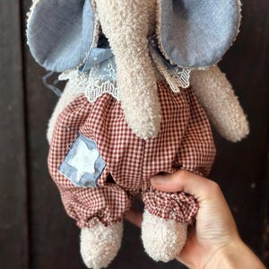 May include: A plush elephant doll with a cream-colored body, blue denim ears, and red and white checkered pants. The doll has a star-shaped patch on the pants and a lace collar. A charming handmade toy.