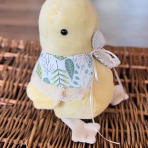 May include: A plush yellow chick with a decorative bib featuring a green and white leaf pattern and lace trim. The chick has a small black eye and a white bow. The chick is sitting on a woven basket.