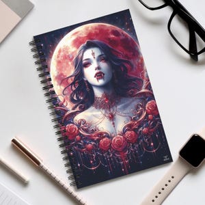 Könnte beinhalten: Ein spiralgebundenes Notizbuch mit einem dunklen Hintergrund, auf dem eine Frau mit langen schwarzen Haaren und roten Augen zu sehen ist. Sie trägt eine silberne Halskette und eine rote Blumenkrone. Der Hintergrund ist dunkelblau mit einem roten Mond und Sternen.