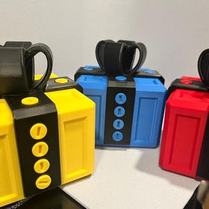 May include: Three colorful boxes in yellow, blue, and red, each with a black handle and accents. The boxes have a rectangular shape with rounded edges and a series of circular details on the front.