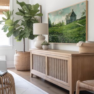 May include: A light wood cabinet with two doors featuring a woven design. The cabinet is sitting on a light wood floor. A large green plant is in a woven basket to the left of the cabinet. A framed painting of a house in a field of flowers is hanging on the wall above the cabinet.