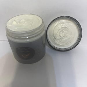 Op de afbeelding: Twee open potten witte crème. Eén pot staat rechtop, de andere ligt op zijn kant. De crème heeft een dikke, opgeklopte textuur. De potten zijn helder met donkergrijze etiketten. De etiketten hebben tekst en een bananenafbeelding.