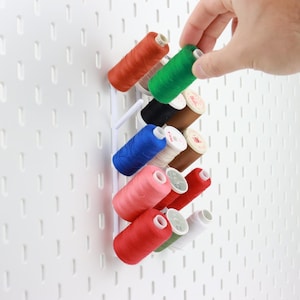 Könnte beinhalten: Eine weiße Lochwand mit einem weißen Garnrollenhalter. Der Halter ist mit Garnrollen in verschiedenen Farben gefüllt, darunter Rot, Grün, Blau und Rosa. Eine Hand greift nach einer grünen Garnrolle.