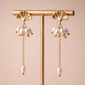 May include: Gold-toned earrings with a fan-shaped top and dangling pearls. The earrings are displayed on a gold stand against a soft pink background. The design features small gold beads and a delicate chain.