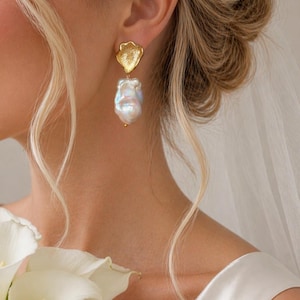 Pode incluir: Brincos pendurados dourados com um design floral texturizado e uma grande pérola irregular. A pessoa usa um vestido branco e segura um buquê de copo-de-leite branco. Joias de noiva.