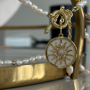 以下が含まれることがあります： パールチェーンとペンダントが付いたゴールドトーンのネックレス。ペンダントは白い背景にサンバーストのデザインと小さな輝く石が施されています。ネックレスは金色のスタンドに展示されています。