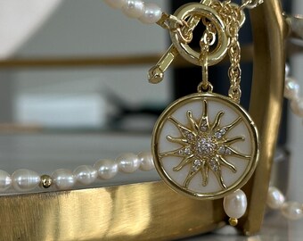 Peut inclure: Un collier doré avec une chaîne de perles et un pendentif. Le pendentif a un fond blanc avec un motif de soleil et de petites pierres scintillantes. Le collier est présenté sur un support doré.