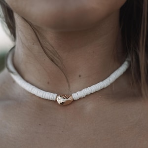 以下が含まれることがあります： 白い貝殻のネックレスに金のペンダントが付いている。ネックレスは、細い紐に小さな丸い貝殻が繋がっている。ペンダントは、金色の仕上げが施された小さな楕円形の貝殻である。